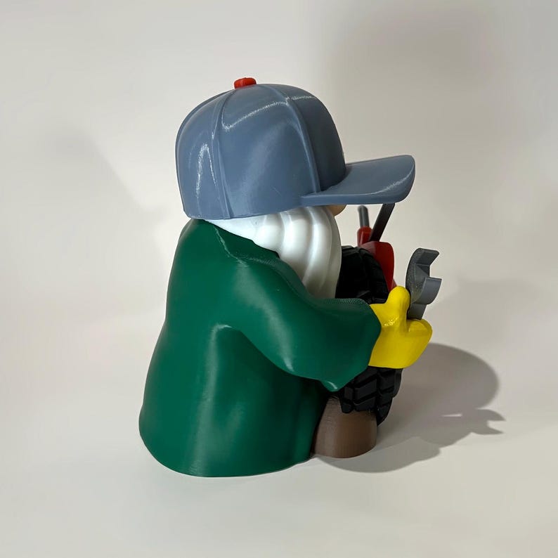 May include: A 3D-printed figurine of a mechanic gnome wearing a gray cap with a red button, a green jacket, and yellow gloves. The gnome holds a wrench and appears to be working on a small vehicle. The figurine has a white beard.