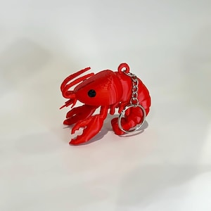 May include: A bright red lobster keychain with a silver chain and keyring. The lobster has detailed claws, antennae, and a textured body. The keychain is a fun accessory.