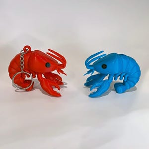 May include: Two lobster ornaments, one red and one blue, are shown against a white backdrop. The red lobster has a keyring attached. Both have detailed claws, antennae, and eyes. The blue lobster is a solid colour, while the red lobster has a slightly textured appearance.