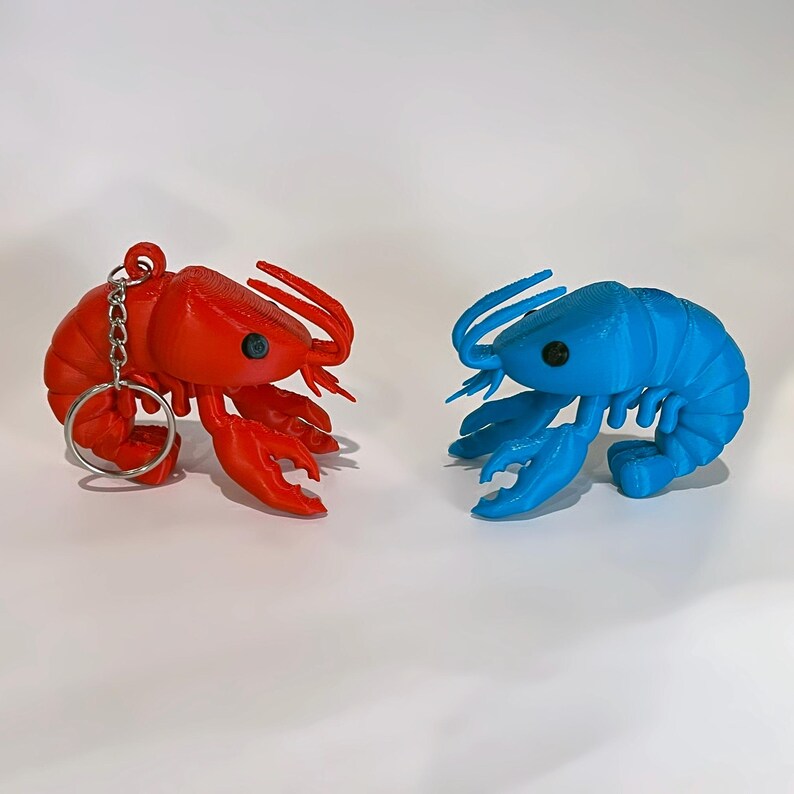 May include: Two lobster ornaments, one red and one blue, are shown against a white backdrop. The red lobster has a keyring attached. Both have detailed claws, antennae, and eyes. The blue lobster is a solid colour, while the red lobster has a slightly textured appearance.