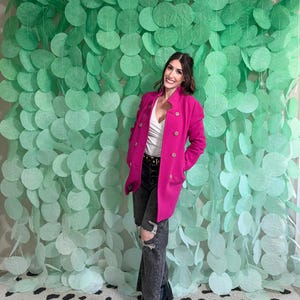 May include: A woman wearing a bright pink double-breasted coat with gold buttons, black jeans with ripped knees, and black boots. The background features a green, circular paper decoration and a black and white patterned rug.