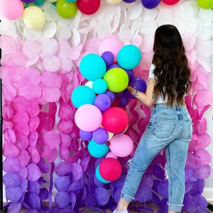 May include: A colorful balloon arch with pink, purple, green, blue, and yellow balloons. The arch is in front of a wall decorated with paper circles in shades of pink, purple, and white.