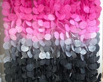 May include: A decorative wall hanging with a colour gradient of pink, silver, and black circular paper cutouts. The circles are strung together, creating a layered effect. The background is a white wall.