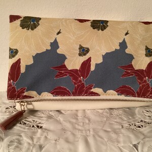 May include: A clutch purse with a zipper closure and a tassel. The purse is made of a cream-colored fabric with a floral pattern in shades of red, blue, and green.
