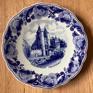 May include: A blue and white ceramic plate with a decorative floral border. The center of the plate features a depiction of a castle with the text "Maastricht Helpoort".