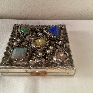 May include: A silver compact with an ornate design featuring colorful stones and faux pearls. The compact has a hinged lid and a small gold latch.
