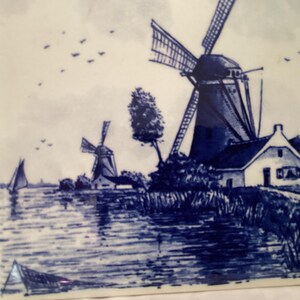 May include: A blue and white ceramic tile depicting a Dutch landscape with windmills, a house, and a canal. The tile is painted in a traditional Delft blue style.