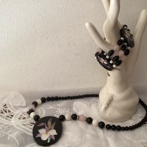 May include: A black and white necklace with a floral pendant. The pendant is made of black and white shell and has a pink flower in the center. The necklace is made of black and white beads.