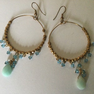 May include: Gold hoop earrings with a teardrop-shaped turquoise stone dangling from each hoop. The hoops are adorned with small blue crystals.