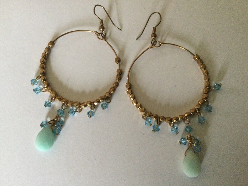 May include: Gold hoop earrings with a teardrop-shaped turquoise stone dangling from each hoop. The hoops are adorned with small blue crystals.