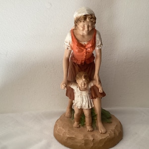 May include: A ceramic figurine of a woman in a red and white dress holding a small child in a white dress. The figurine is standing on a brown base.