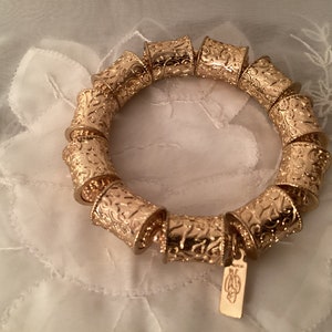 May include: Gold bracelet with a decorative, ornate design. The bracelet is made of multiple linked pieces, each with a unique pattern. The bracelet has a small tag attached with the brand name.