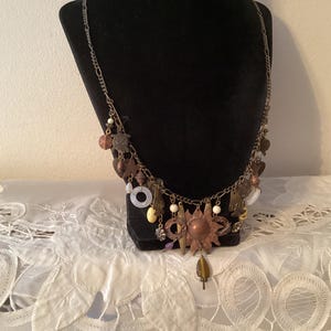 May include: A vintage-style necklace with a variety of charms and pendants. The necklace features a copper sunburst pendant, along with various metal, bead, and stone accents. The chain is a silver color, and the necklace is displayed on a black bust.