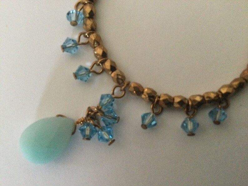 May include: A gold beaded necklace with a light blue teardrop pendant and blue crystal accents.