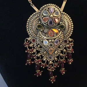 May include: A gold necklace with a large, ornate pendant featuring a circular design with multi-colored gemstones and a smaller, crescent-shaped design with more gemstones. The pendant is attached to a chain with a series of dangling burgundy beads.