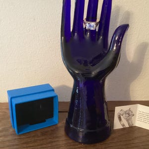 May include: A blue glass hand ring holder with a gold ring with a large clear square stone. The ring is on the index finger of the hand. A blue box is on the table next to the hand. A small card with text is on the table.