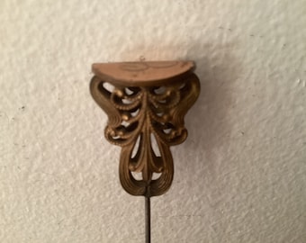 May include: An antique hat pin with a decorative brass head. The head features an ornate, stylized design with a half-circle top and a long, slender pin. The pin is likely used for securing hats or other accessories.