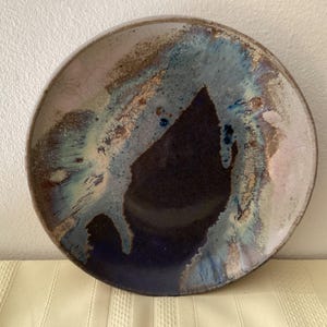 May include: A round ceramic plate featuring a deep indigo center that transitions into swirling patterns of light blue, cream, and earthy brown tones. The glaze has a reactive finish with visible texture and a glossy sheen across the surface.