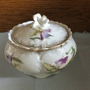May include: A white porcelain trinket box with a lid. The box is decorated with hand-painted purple, yellow, and green flowers. The lid has a raised flower knob.