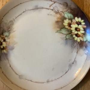 May include: A white porcelain plate with a gold rim and a delicate floral design in shades of yellow, green, and brown. The flowers are painted in a realistic style and appear to be daisies.