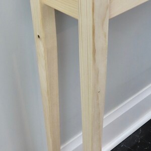 Puede incluir: Una mesa consola de madera clara con un dise&ntilde;o sencillo. La mesa tiene una tapa rectangular y cuatro patas. Las patas son c&oacute;nicas y tienen una forma ligeramente curvada. La mesa est&aacute; hecha de madera sin tratar y tiene una veta de madera natural.