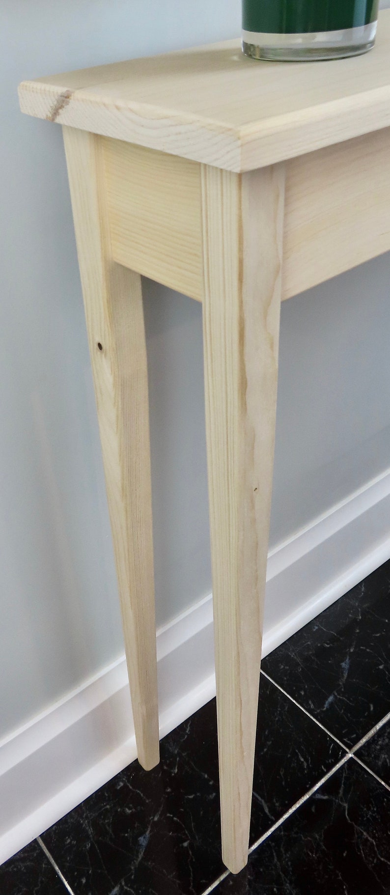 Puede incluir: Una mesa consola de madera clara con un dise&ntilde;o sencillo. La mesa tiene una tapa rectangular y cuatro patas. Las patas son c&oacute;nicas y tienen una forma ligeramente curvada. La mesa est&aacute; hecha de madera sin tratar y tiene una veta de madera natural.