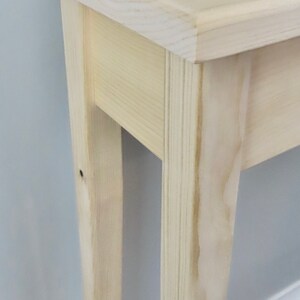 Puede incluir: Primer plano de una mesa consola de madera clara con un dise&ntilde;o sencillo. La mesa tiene una tapa rectangular y dos patas a cada lado. La veta de la madera es visible en la superficie de la mesa.