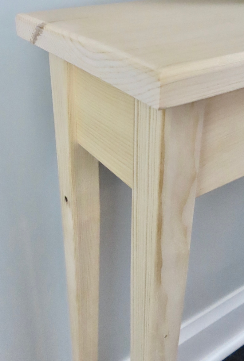Puede incluir: Primer plano de una mesa consola de madera clara con un dise&ntilde;o sencillo. La mesa tiene una tapa rectangular y dos patas a cada lado. La veta de la madera es visible en la superficie de la mesa.