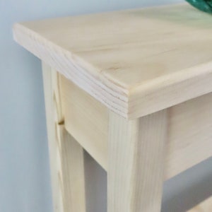 May include: A light wood console table with a simple design. The table has a rectangular top and two shelves. The table is made of unfinished wood.