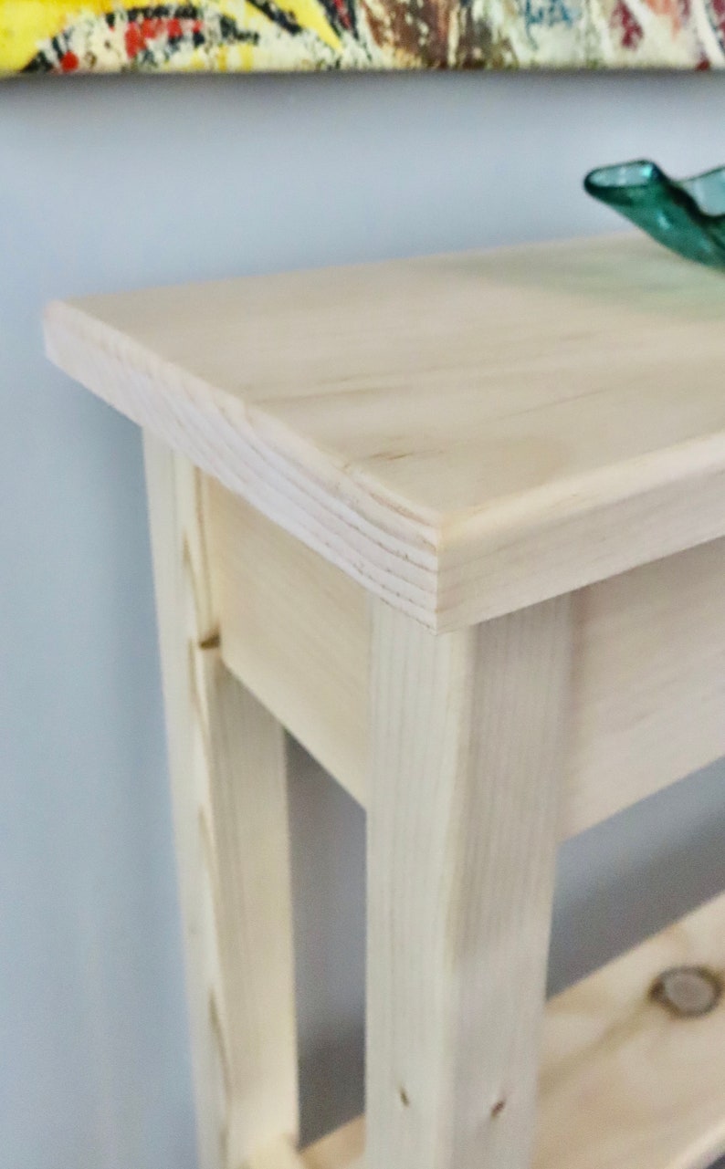 May include: A light wood console table with a simple design. The table has a rectangular top and two shelves. The table is made of unfinished wood.