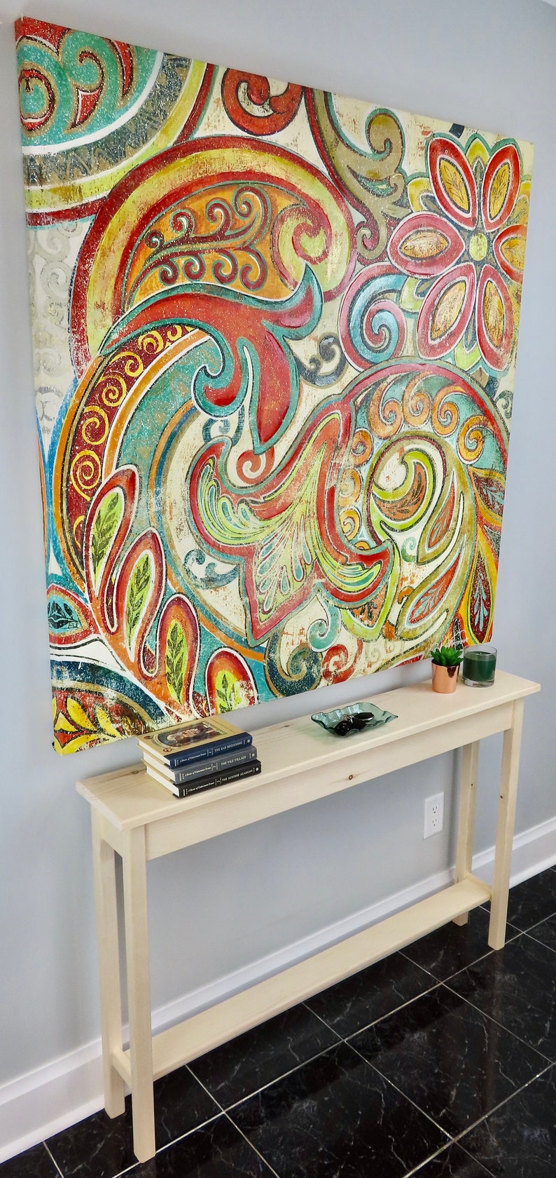 May include: A light wood console table with a shelf, featuring a colorful abstract painting with floral motifs hanging above it. The table is decorated with a small potted plant, a glass dish with stones, and a stack of books.
