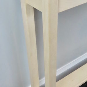 May include: A white wooden console table with a single shelf. The table has a simple design with straight lines and a rectangular top. The table is standing on a black and white tiled floor.