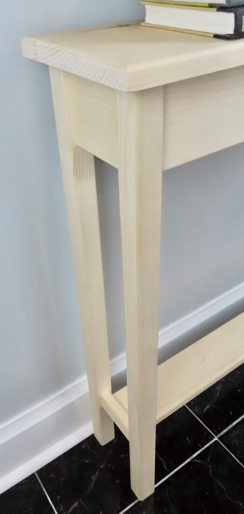 May include: A white wooden console table with a single shelf. The table has a simple design with straight lines and a rectangular top. The table is standing on a black and white tiled floor.