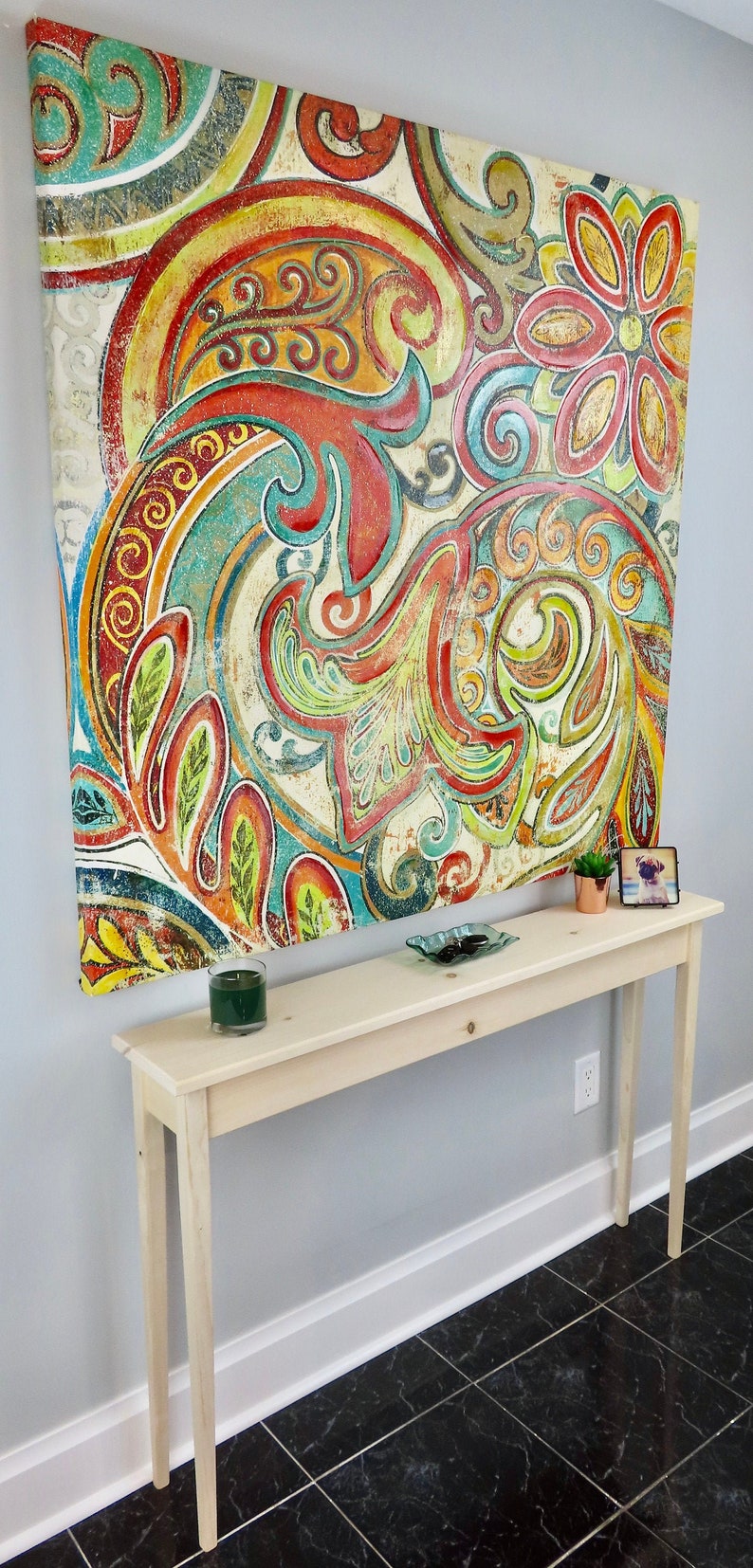 Puede incluir: Una mesa consola de madera clara con una pintura decorativa de lienzo floral colgada encima. La mesa tiene un dise&ntilde;o sencillo con dos patas a cada lado y una parte superior plana. La pintura presenta un patr&oacute;n paisley colorido con tonos rojos, naranjas, amarillos, verdes y azules.