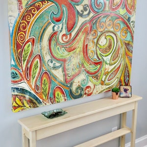May include: A light wood console table with two shelves. The table is in front of a wall with a large abstract painting featuring colorful paisley patterns.