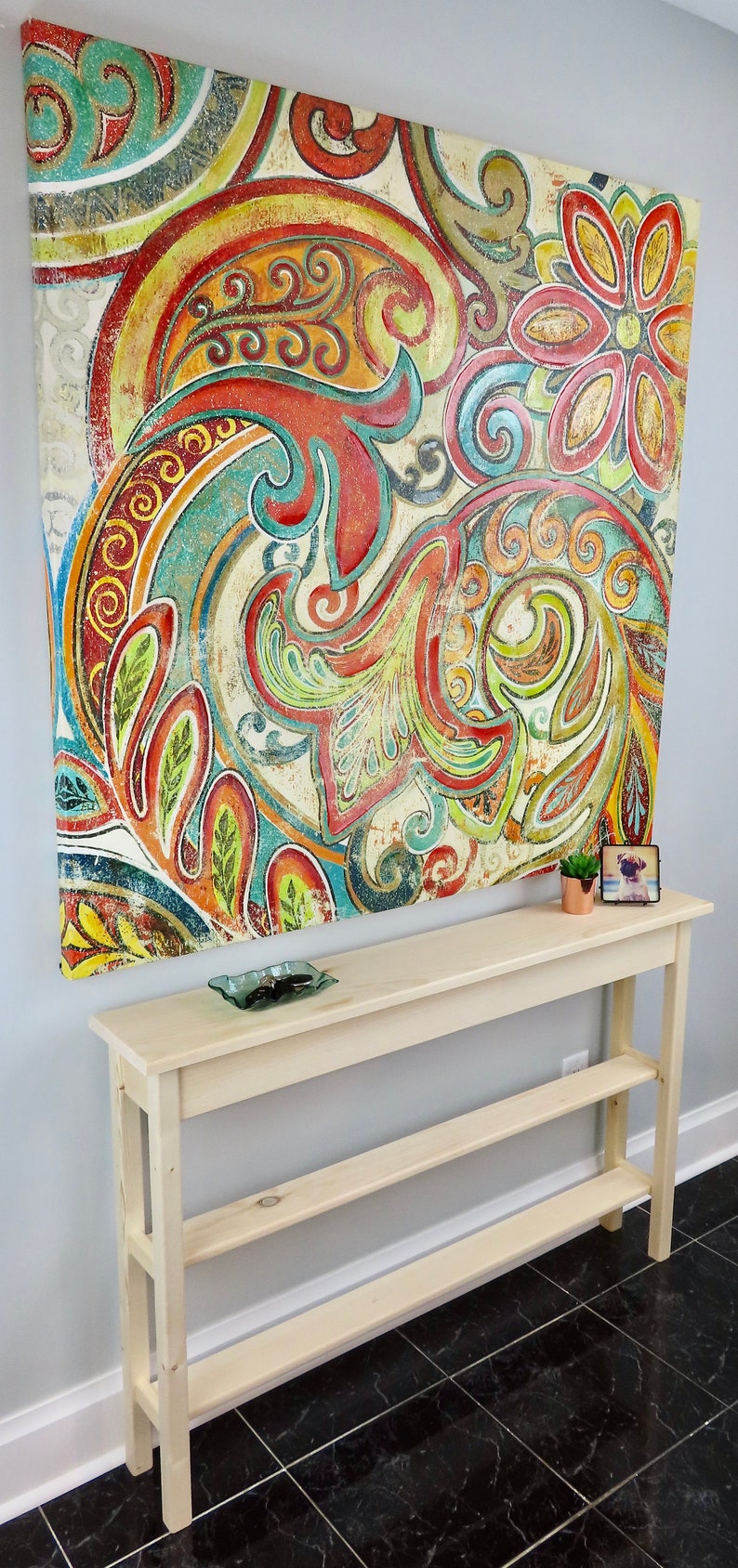 May include: A light wood console table with two shelves. The table is in front of a wall with a large abstract painting featuring colorful paisley patterns.