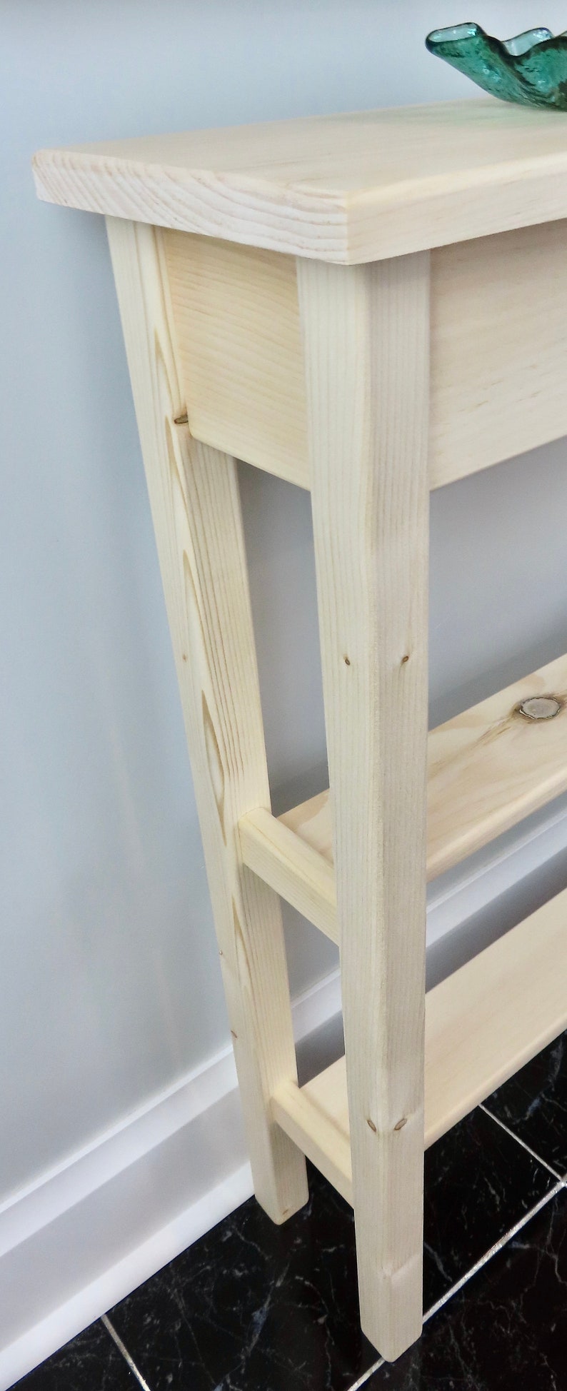 May include: A light wood console table with two shelves. The table has a simple design with straight lines and a natural finish.