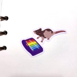 May include: A sticker featuring a brown and tan rodent with a long tail, positioned near a purple xylophone with rainbow-colored keys. The sticker is on a white background with binder rings.