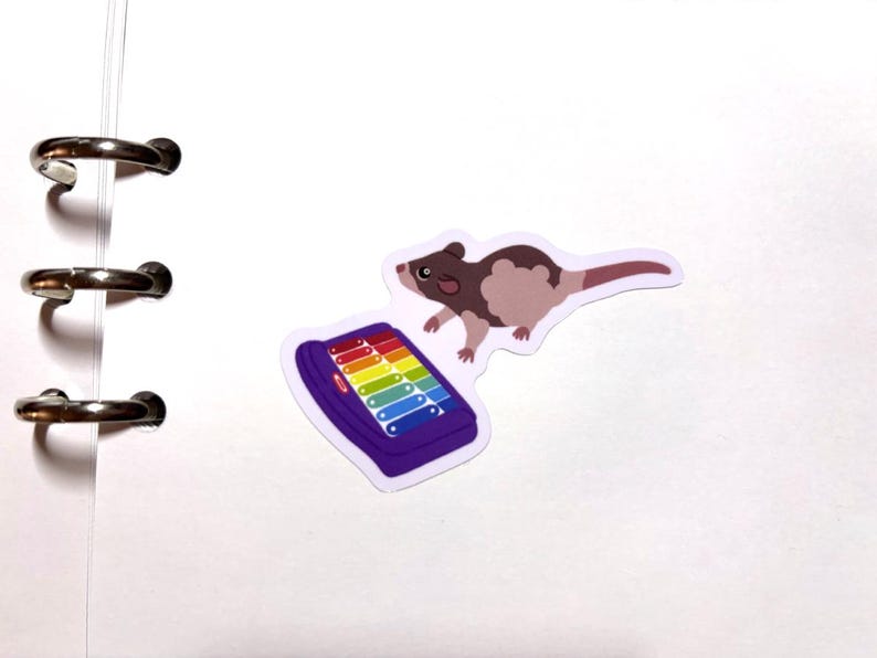 May include: A sticker featuring a brown and tan rodent with a long tail, positioned near a purple xylophone with rainbow-colored keys. The sticker is on a white background with binder rings.