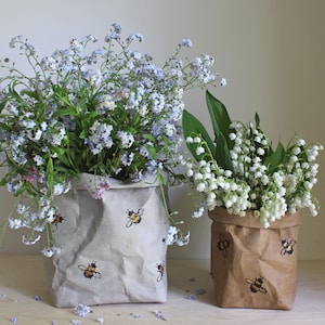 Könnte beinhalten: Zwei Papierbeutel mit zerknitterter Textur, einer braun und einer grau, beide mit Bienen-Illustrationen verziert. Die Beutel sind mit Blumensträußen gefüllt, einer mit blauen Vergissmeinnicht und der andere mit weißen Maiglöckchen.