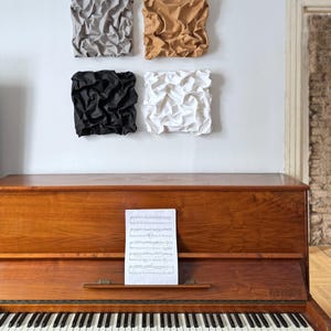 May include: A wooden piano with a sheet of music on the stand, and four square wall art pieces in gray, tan, black, and white. The piano has black and white keys and a polished wood finish.