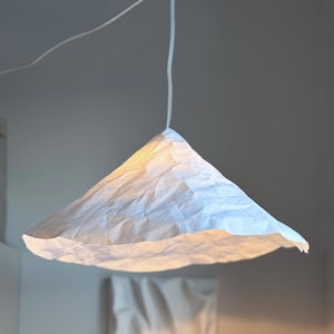Può includere: Una lampada a sospensione bianca a forma di cono realizzata in carta strutturata. La luce è appesa a un cavo bianco e illumina la stanza sottostante. La carta ha un aspetto stropicciato, creando un design unico e moderno.