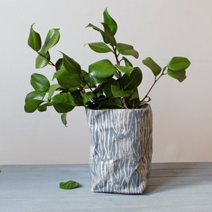 Könnte beinhalten: Eine grau-weiß gemusterte Vase enthält leuchtend grüne, belaubte Zweige. Die Vase hat ein strukturiertes, papierartiges Aussehen. Die Blätter sind glänzend und voll und bilden eine natürliche, dekorative Anordnung.