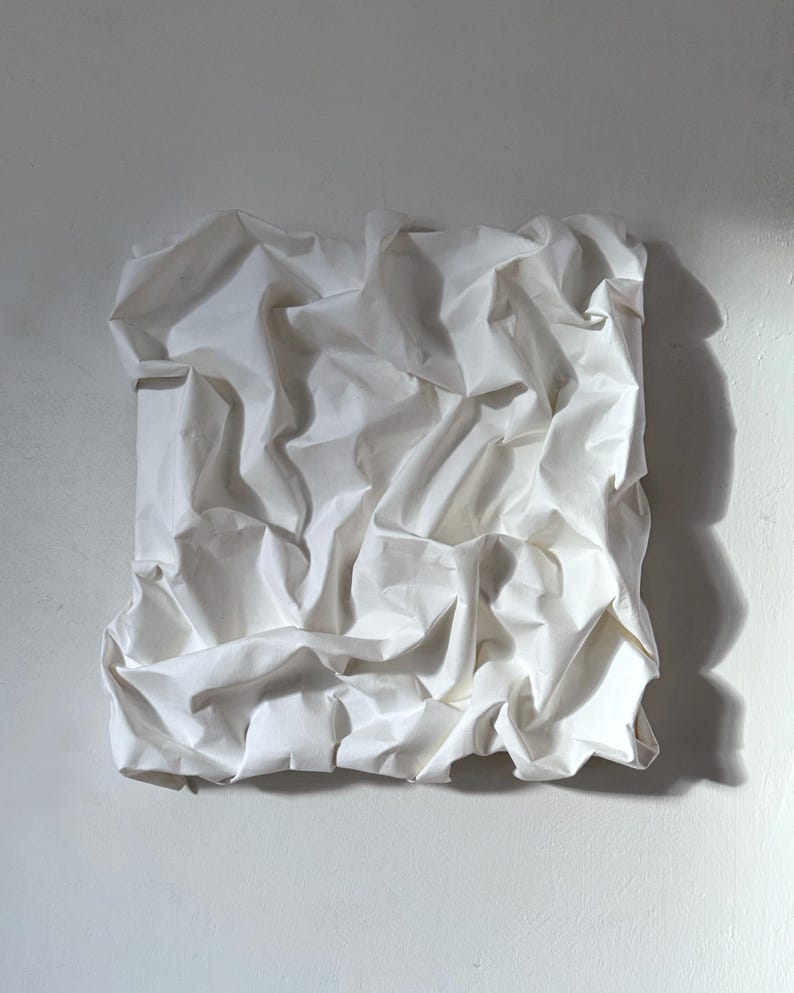 May include: A square, white, crumpled paper sculpture mounted on a white wall. The paper is heavily textured with deep folds and shadows, creating a dynamic and abstract composition. The lighting emphasizes the paper's irregular surface.
