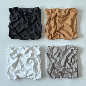 May include: Four square fabric art pieces in black, tan, white, and gray. Each piece has a crumpled, textured appearance, creating a unique visual effect. The art pieces are mounted on a white background, showcasing the colors and textures.