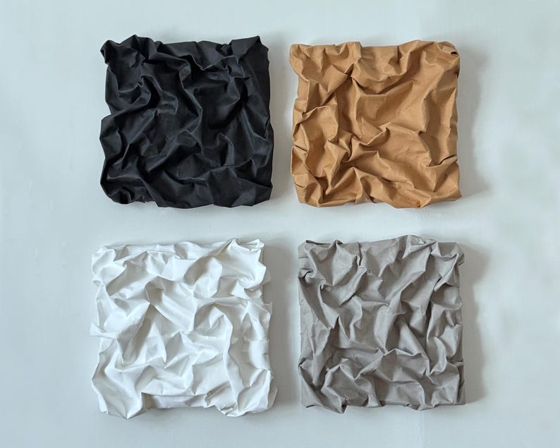 May include: Four square fabric art pieces in black, tan, white, and gray. Each piece has a crumpled, textured appearance, creating a unique visual effect. The art pieces are mounted on a white background, showcasing the colors and textures.