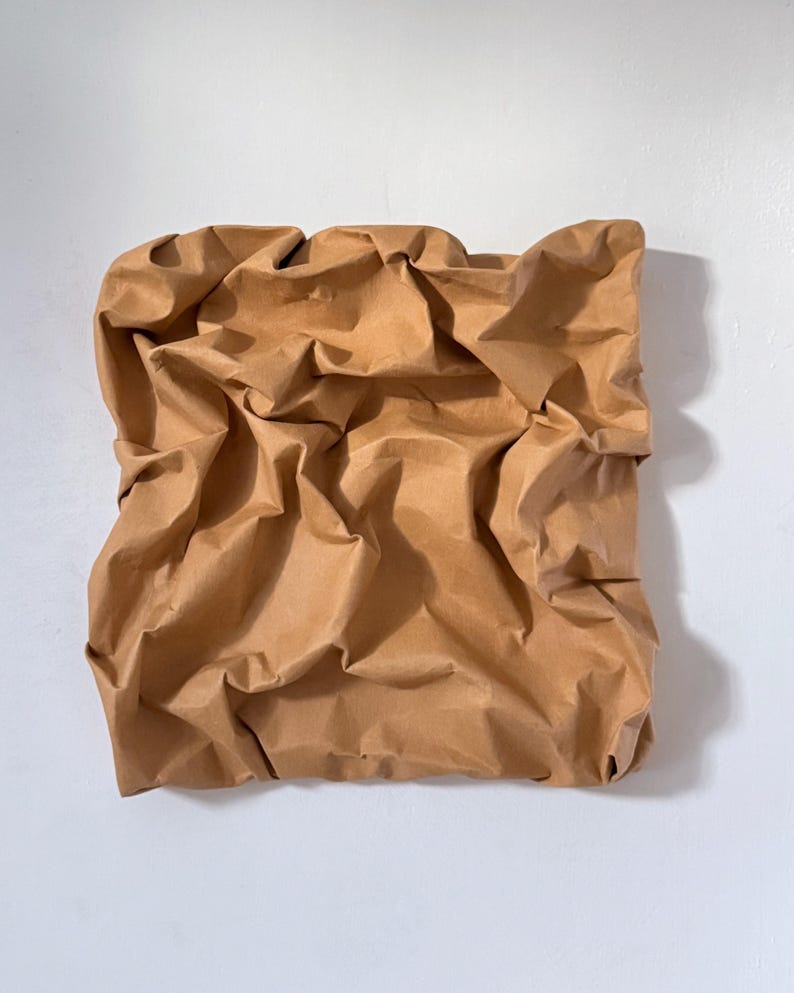 May include: A square, brown paper sculpture with a crumpled texture. The paper is a warm, neutral tone, and the surface is heavily creased, creating shadows and highlights. The artwork is mounted on a white wall.