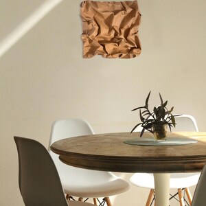 May include: A brown, textured paper art piece is mounted on a beige wall. Below, a round wooden table is surrounded by white and gray chairs. A small potted plant sits on the table.