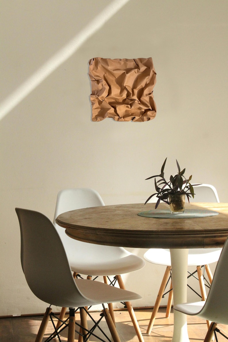 May include: A brown, textured paper art piece is mounted on a beige wall. Below, a round wooden table is surrounded by white and gray chairs. A small potted plant sits on the table.