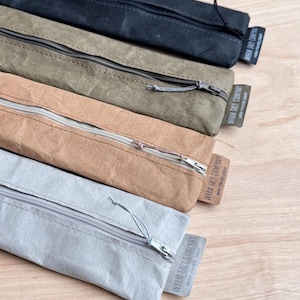 May include: Four rectangular pencil cases in black, olive green, tan, and light gray. Each case features a top zipper closure with a pull cord and a small fabric tag on the side labeled WARM GREY COMPANY water-friendly paper.